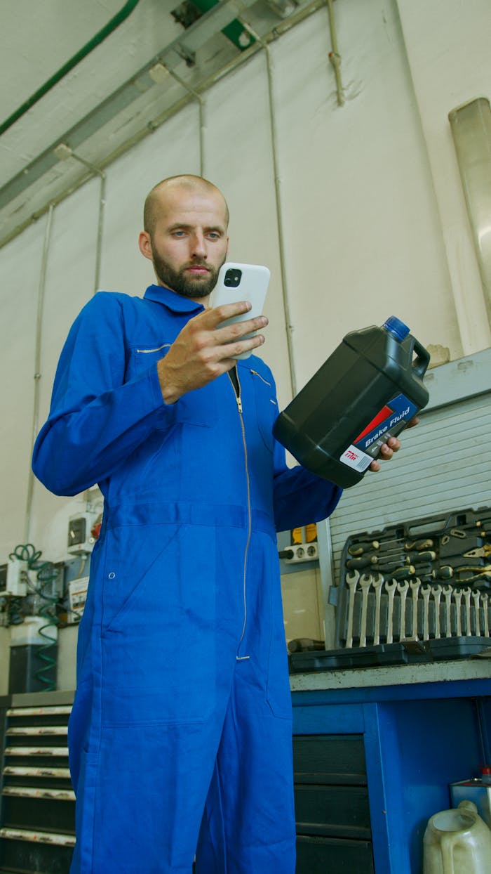 Mechanic in blue coveralls checking brake fluid container while using a smartphone in a garage workshop.