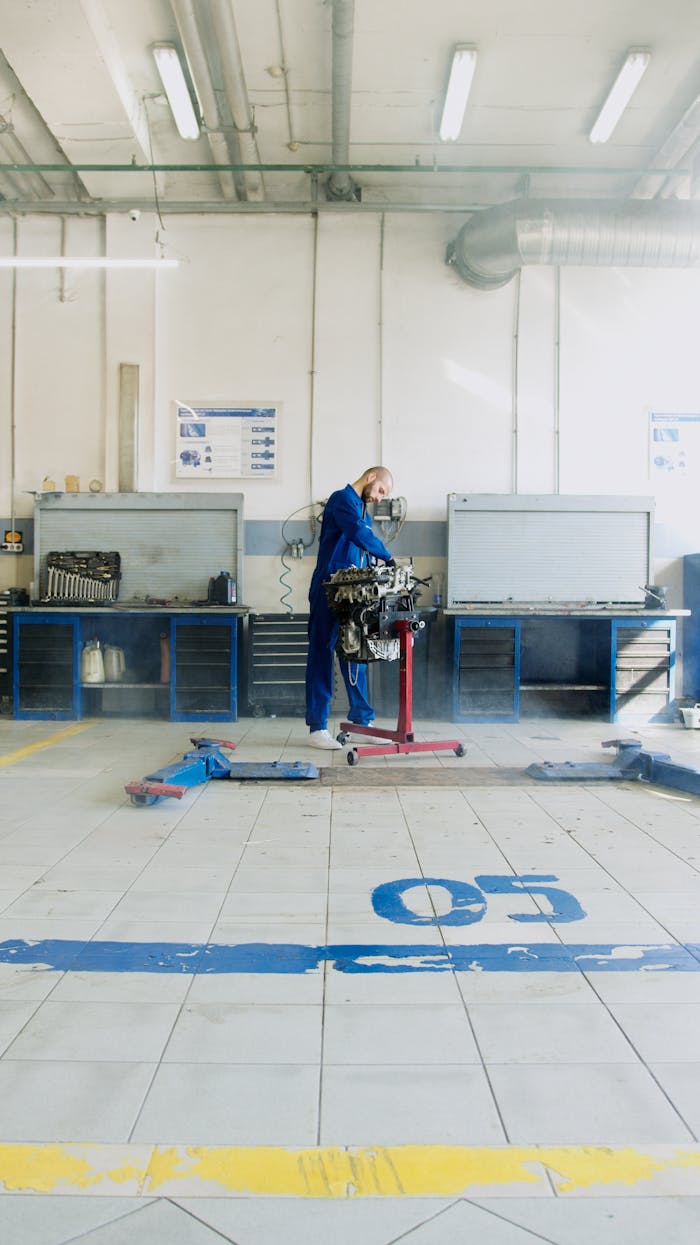 A mechanic in blue overalls repairs an engine in a spacious, well-lit garage.